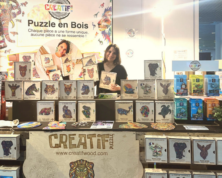 A product display booth for 'CREATIF' wooden puzzles at an exhibition, featuring various wooden puzzle boxes and completed puzzle pieces arranged on shelves and counters, with two women holding up some of the wooden puzzle pieces behind the counter.
