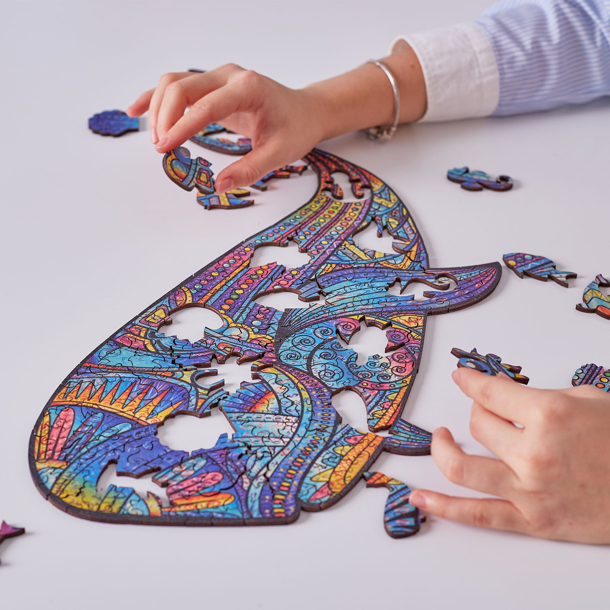 A colorful jigsaw puzzle featuring an intricate whale design, being assembled by two hands on a clean white surface.